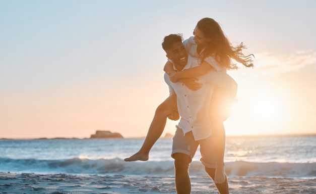 Ein Paar läuft am Strand bei Sonnenuntergang; der Mann trägt die Frau auf dem Rücken.