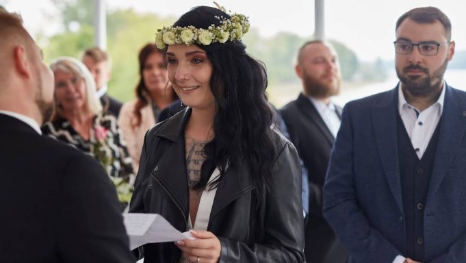 Frau mit Blumenkranz lächelt und spricht während einer Zeremonie, andere Gäste im Hintergrund.